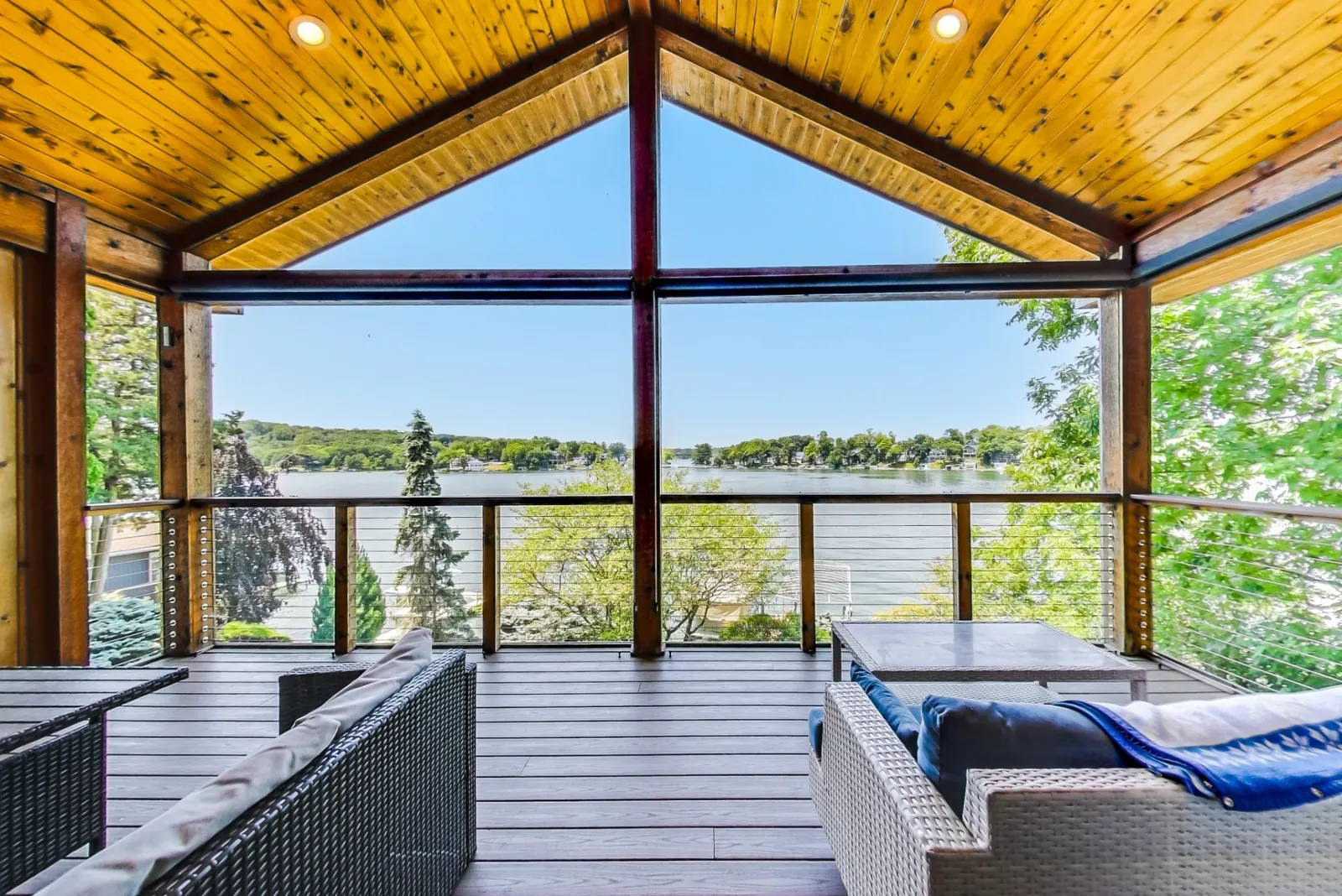 Covered lakefront deck with a vaulted wood ceiling, wicker seating, and cable railings overlooking a Lake Geneva area lake lined with docks and forested shoreline