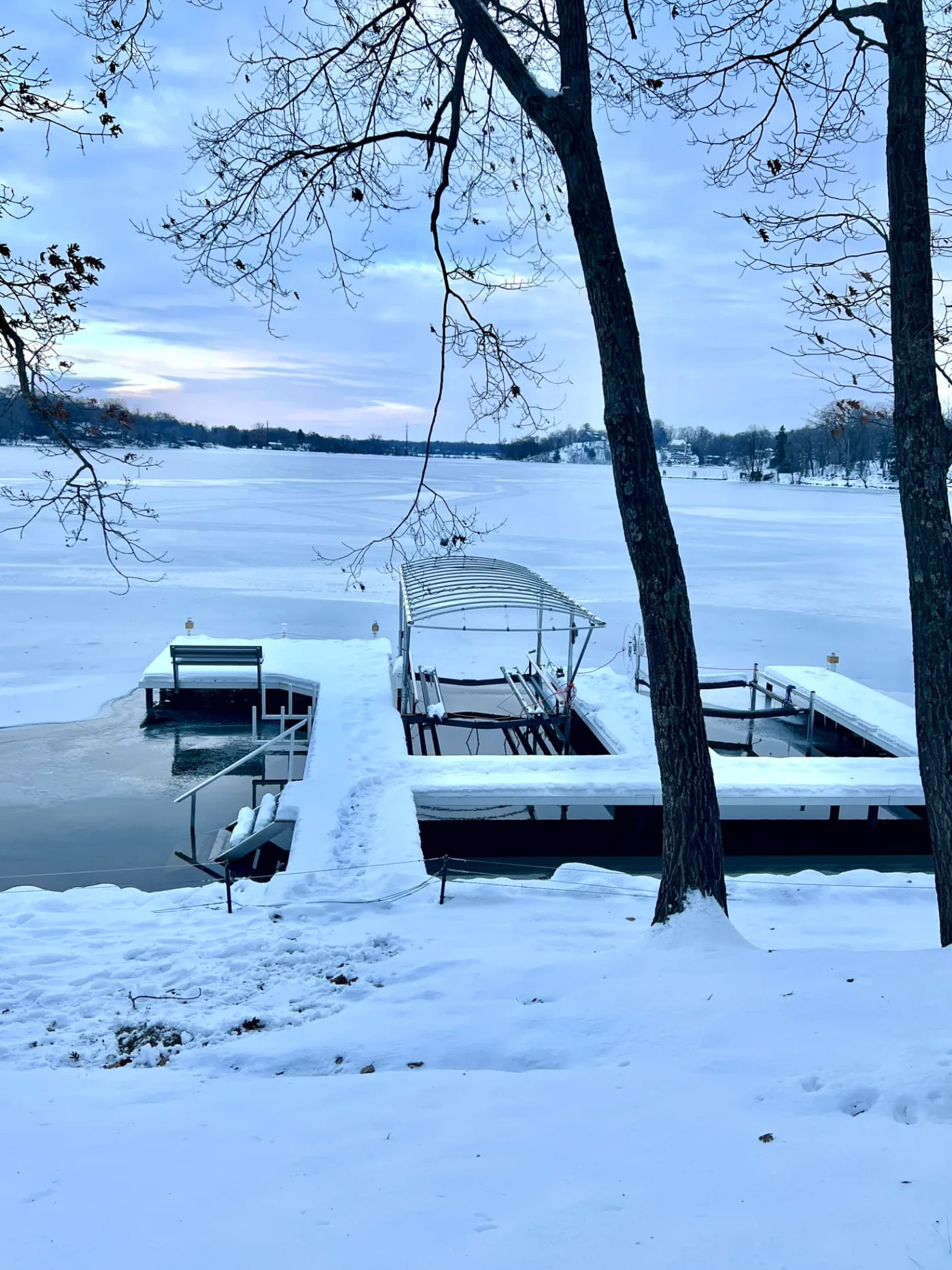 Snow-covered dock and empty boat lift on a frozen Wisconsin lake at winter dawn, with bare trees and lakefront homes on the far shore